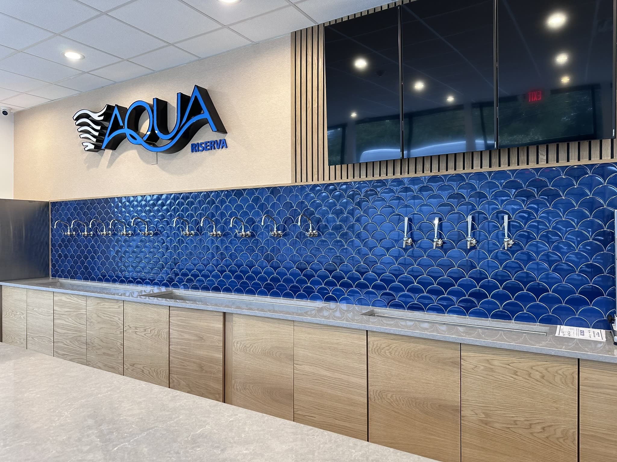 The full Aqua Riserva tap wall in Hawthorne NJ — ten dispensing taps over blue fish-scale tile, stainless trough and custom wood cabinetry beneath